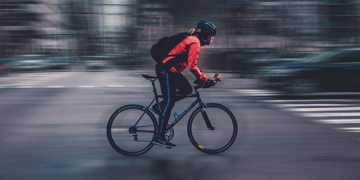 Photo of a cyclist in a black helmet and red jacket on a black and blue racing bike riding through a busy intersection with a blurry backdrop indicating a fast speed.